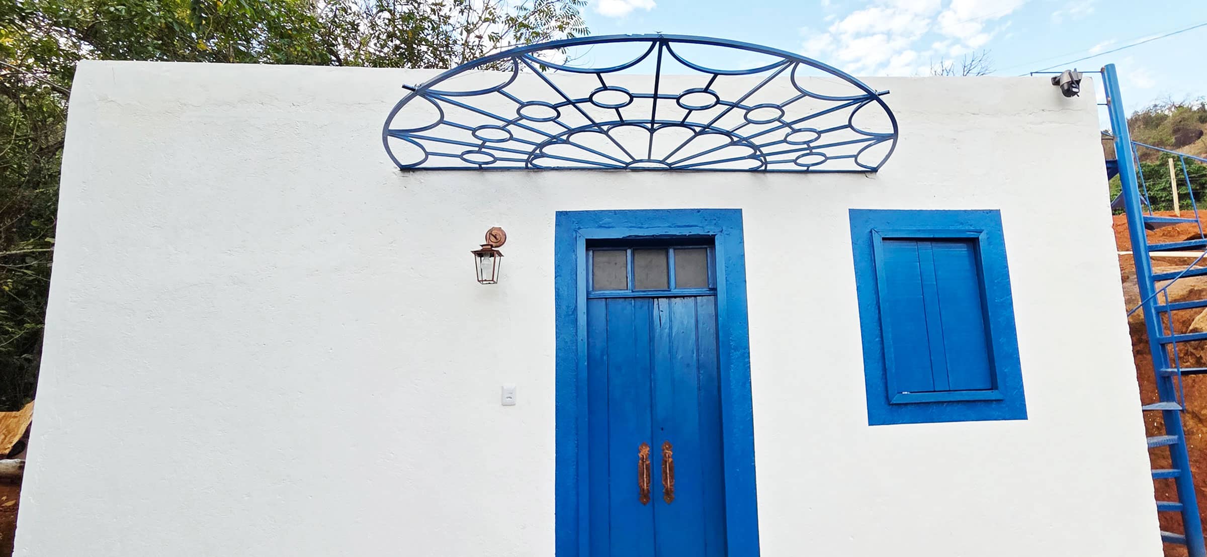 Casa branca com porta e janela azuis sob um gradil de ferro em arco