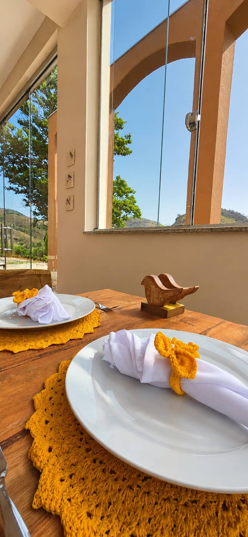 Mesa posta com sousplat de crochê e guardanapo amarelo junto à janela com vista